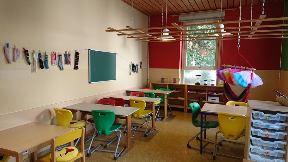 Aufenthaltsraum mit Tischen, farbigen Stühlen und einer Tafel mit Socken auf der Wand 