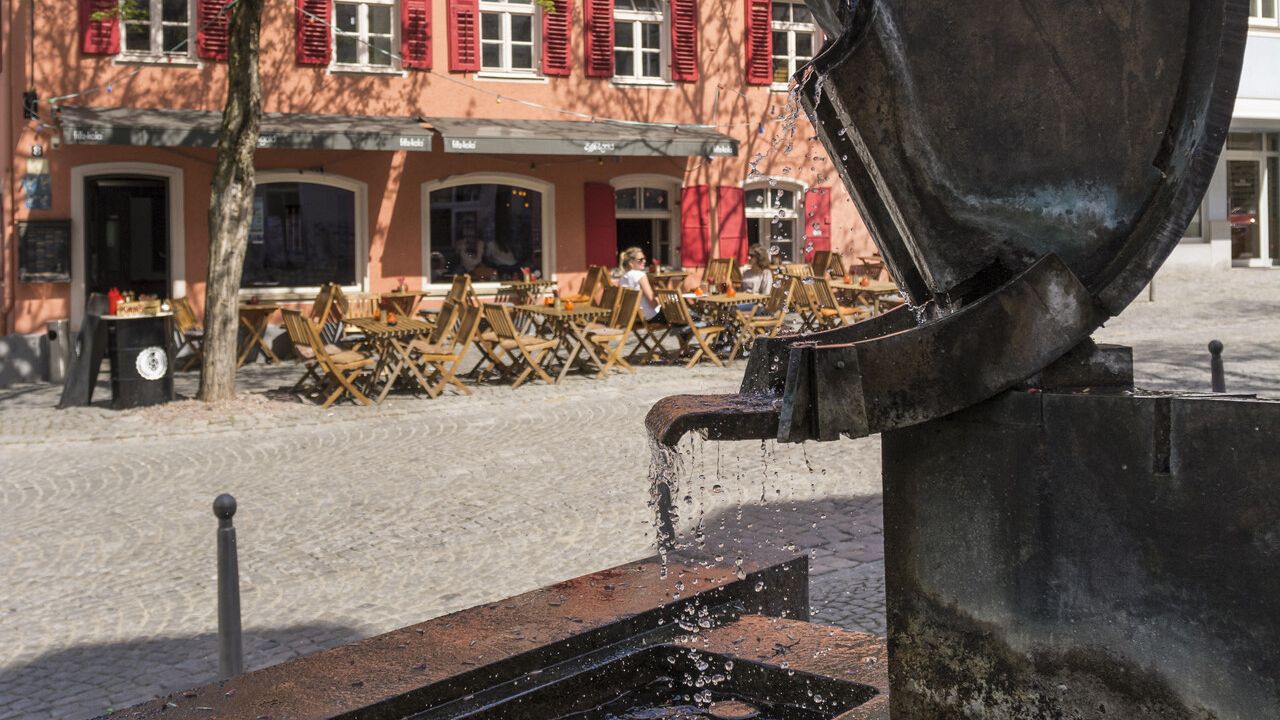 Brunnen, Hans Landner, dahinter ein rötliches Haus mit Gastronomie