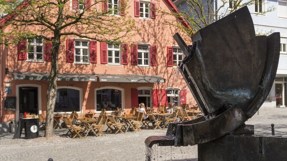 Brunnen, Hans Landner, dahinter ein rötliches Haus mit Gastronomie