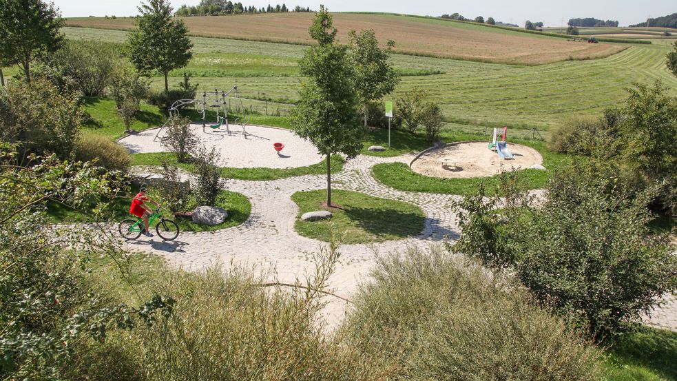 Blick von oben auf den Spielplatz mit zwei Sandkästen, die von Grün umgeben und durch Wege verbunden sind