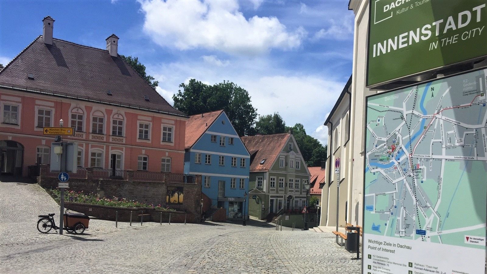 Augsburger Straße in Dachau, linker Hand bunte Häuser des Bezirksmuseums, eines Ladens und des Kochwirts, rechts im Bild eine Informationstafel mit Stadtplan