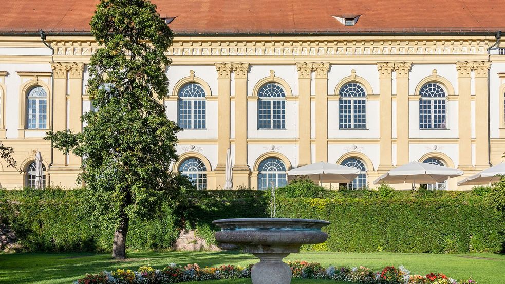 Steinerner Springbrunnen um geben von Blumen und Grün mit Schlossgebäude im Hintergrund