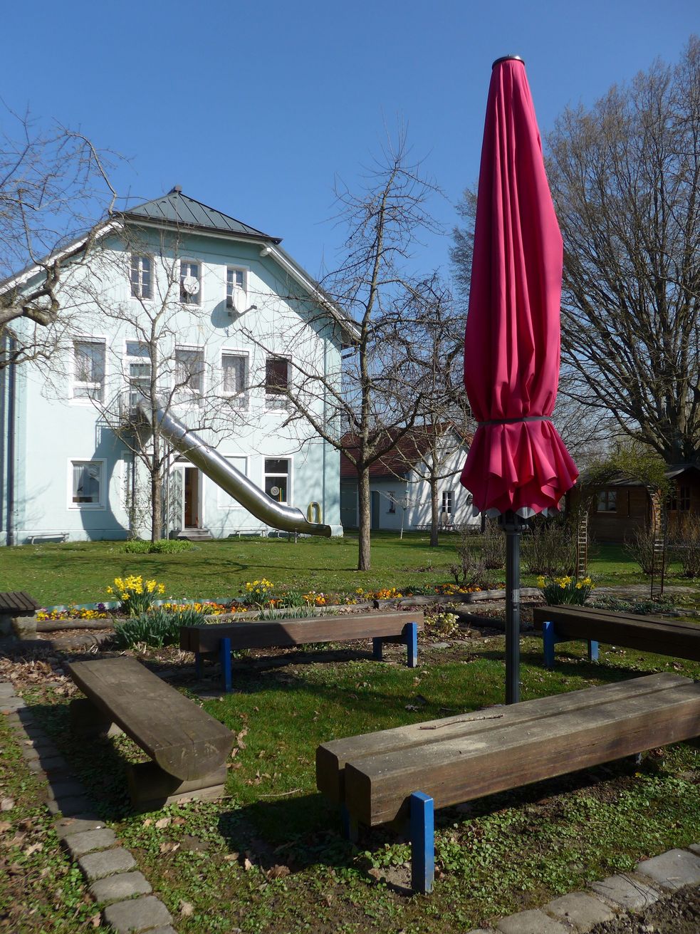 Garten mit Blumen, Bänken um Sonnenschirm, im Hintergrund der Kindergarten mit Rutsche aus dem Gebäude in den Garten