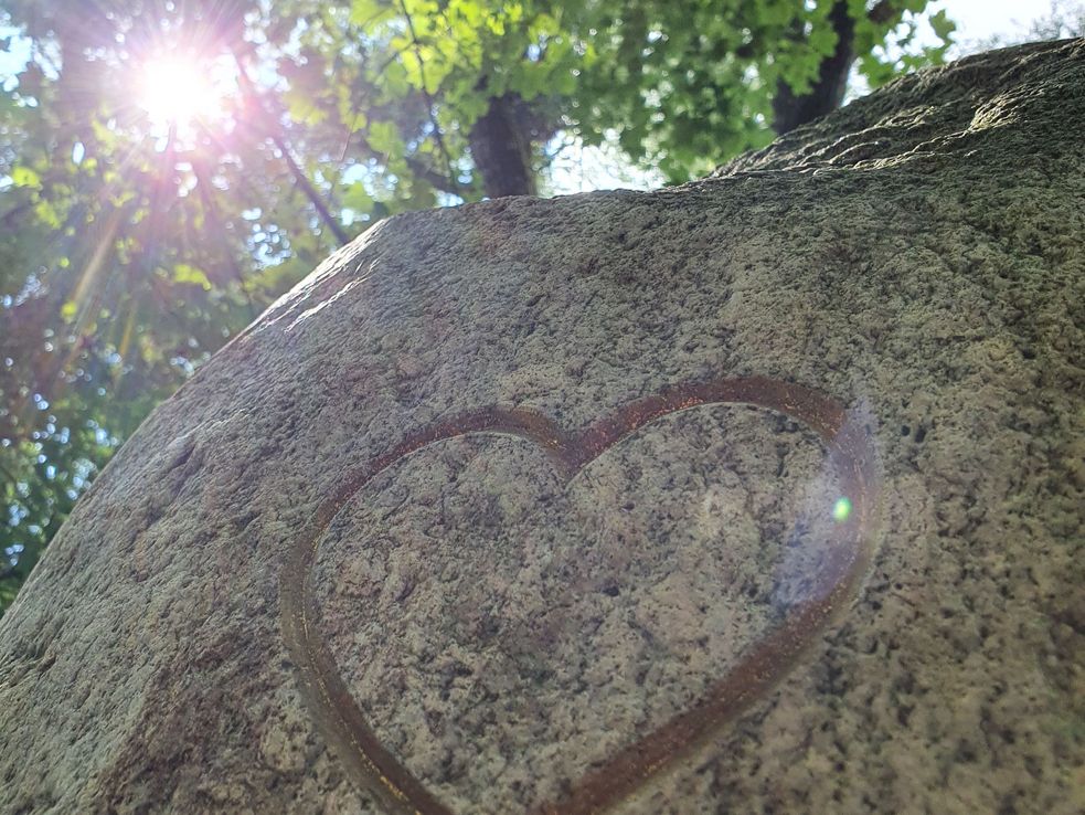 Foto eines Findlings mit goldenem Herz. Im Hintergrund Bäume, durch die die Sonne scheint