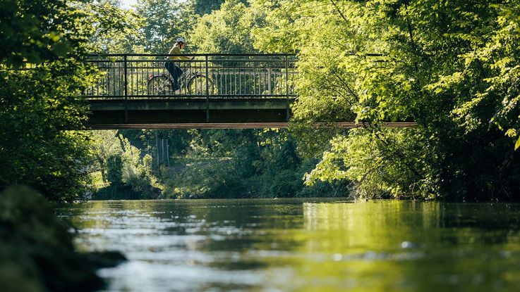 Ein Symbolbild für den Ammer-Amper-Radweg in Dachau: weiblicher Radfahrer auf Brücke, darunter fließt die Amper, umrandet von Bäumen