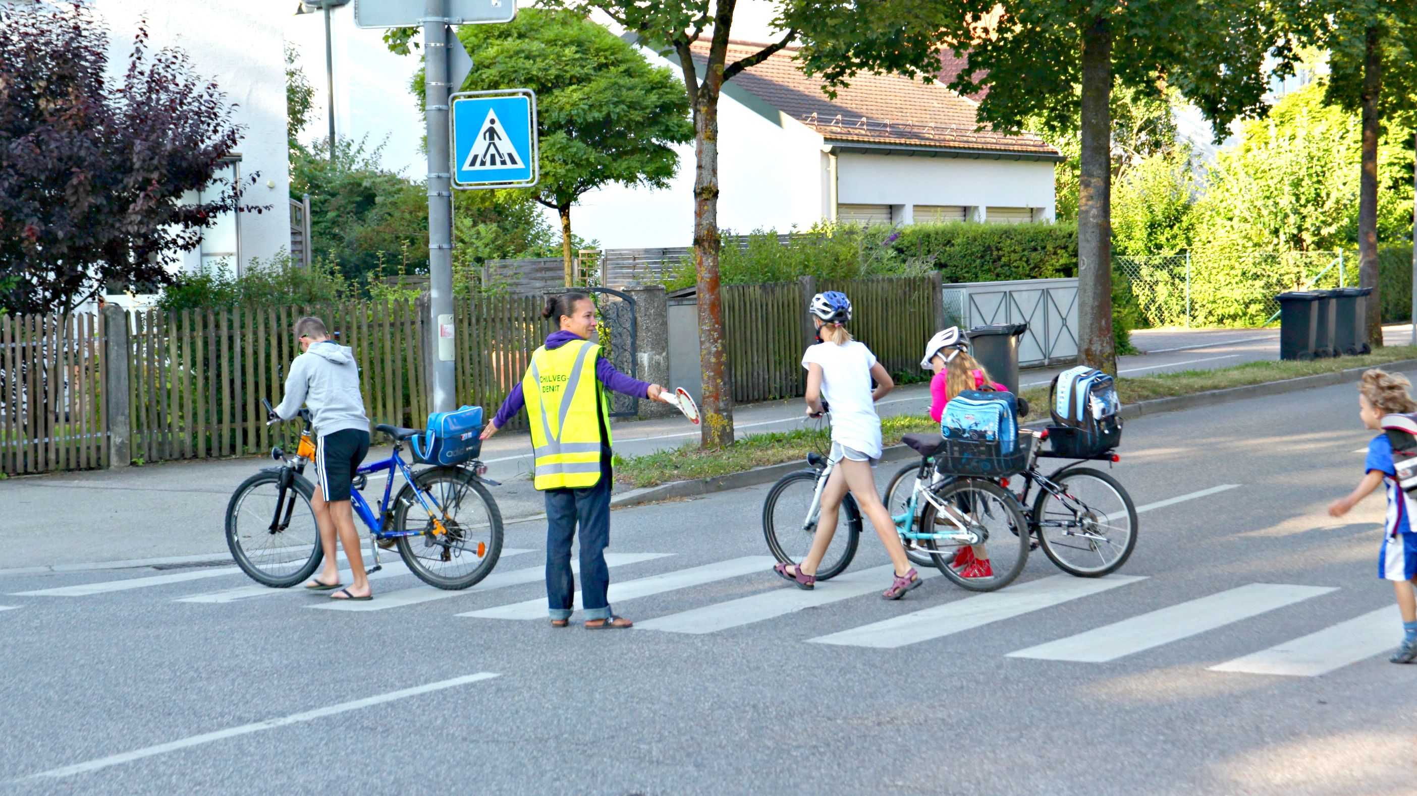 Eine Schulweghelferin mit gelber Warnweste lässt eine Gruppe Schülerinnen und Schüler über einen Zebrastreifen gehen