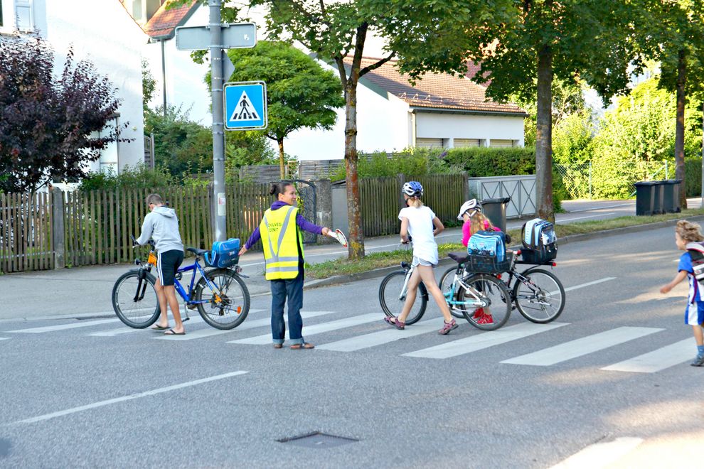 Eine Schulweghelferin mit gelber Warnweste lässt eine Gruppe Schülerinnen und Schüler über einen Zebrastreifen gehen