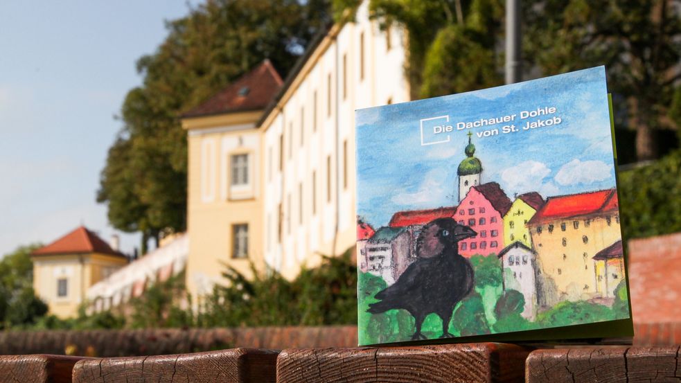 The coolourful children's booklet: "Die Dachauer Dohle von St. Jakob", a souvenir from Dachau photgraphed with a view of the palace buildings and wall in the background. Photo: City of Dachau