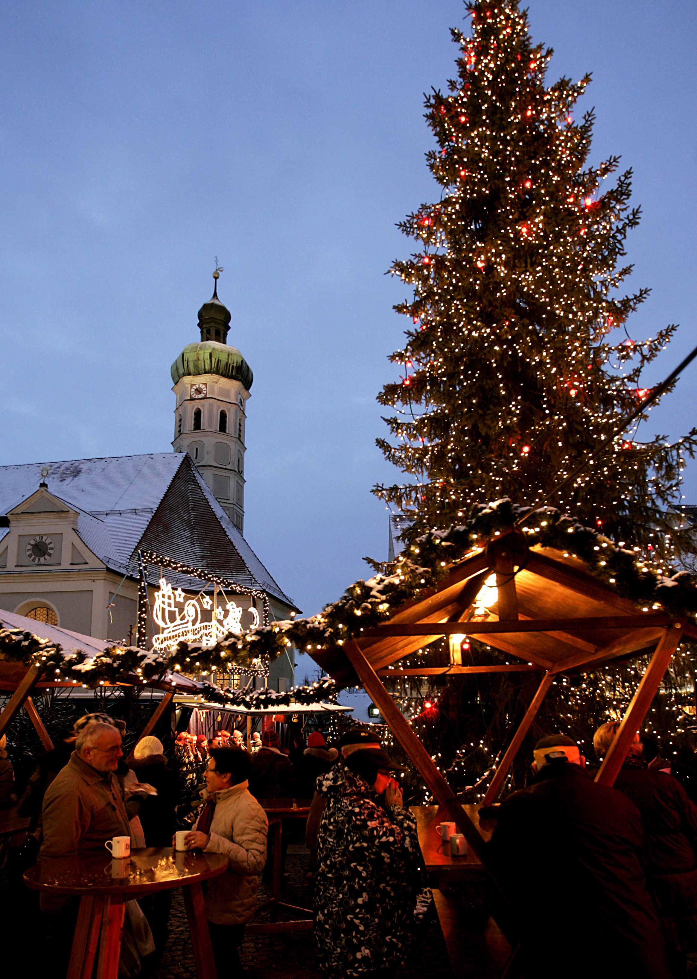 Besucher trinken Glühwein neben beleuchteten Ständen, im Hintergrund Kirche St. Jakob
