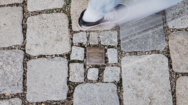 Ein Stolperstein im Pflaster der Dachauer Altstadt und eine Person, die daran vorbeigeht.