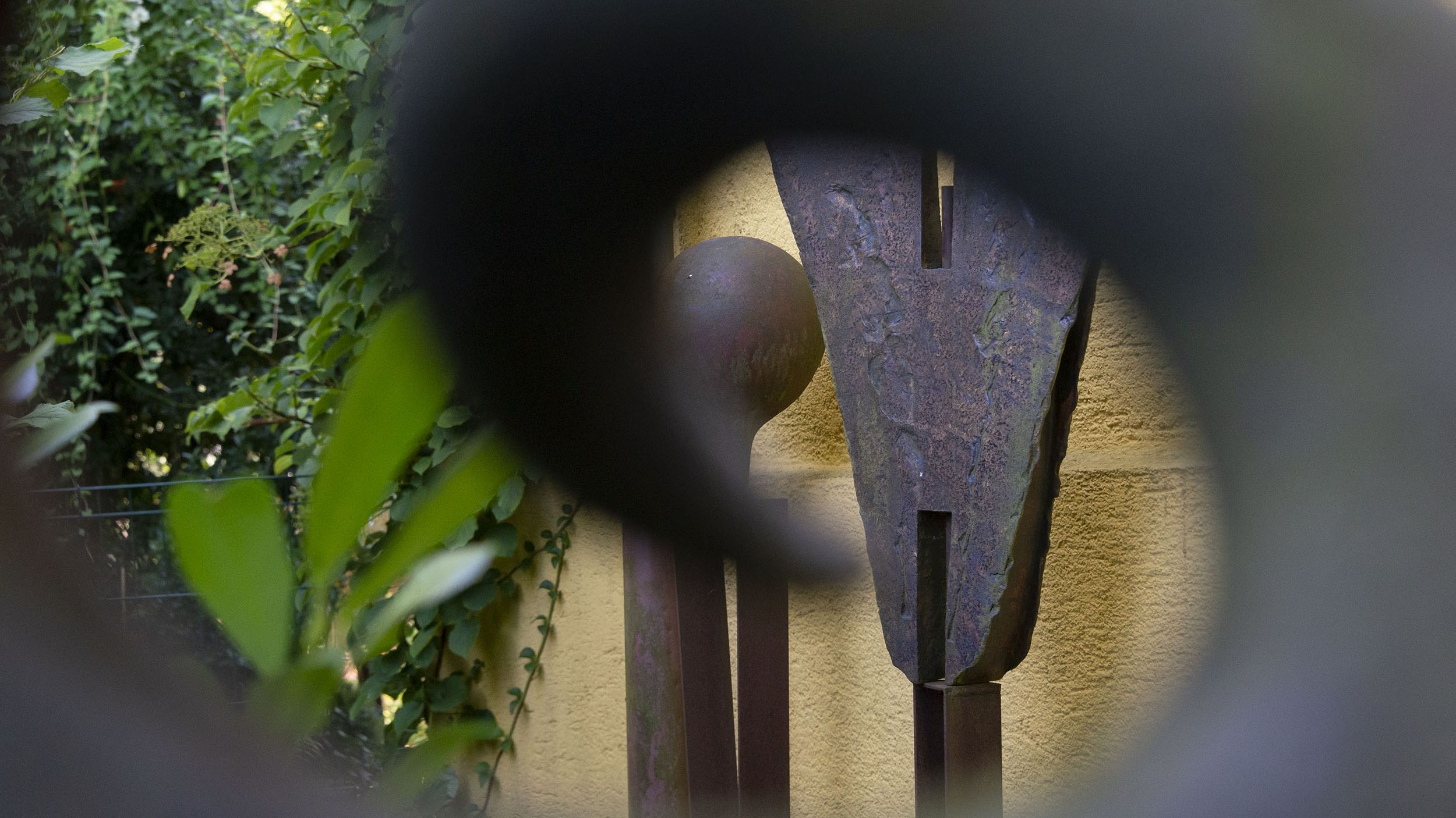 Skulptur von Dachauer Künstlerin Monika Siebmanns, eine Rundung aus Metall gibt den Blick frei auf Skulpturen vor Wand und Pflanzen dahinter.