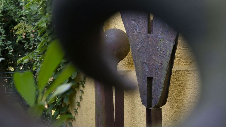 Skulptur von Dachauer Künstlerin Monika Siebmanns, eine Rundung aus Metall gibt den Blick frei auf Skulpturen vor Wand und Pflanzen dahinter.