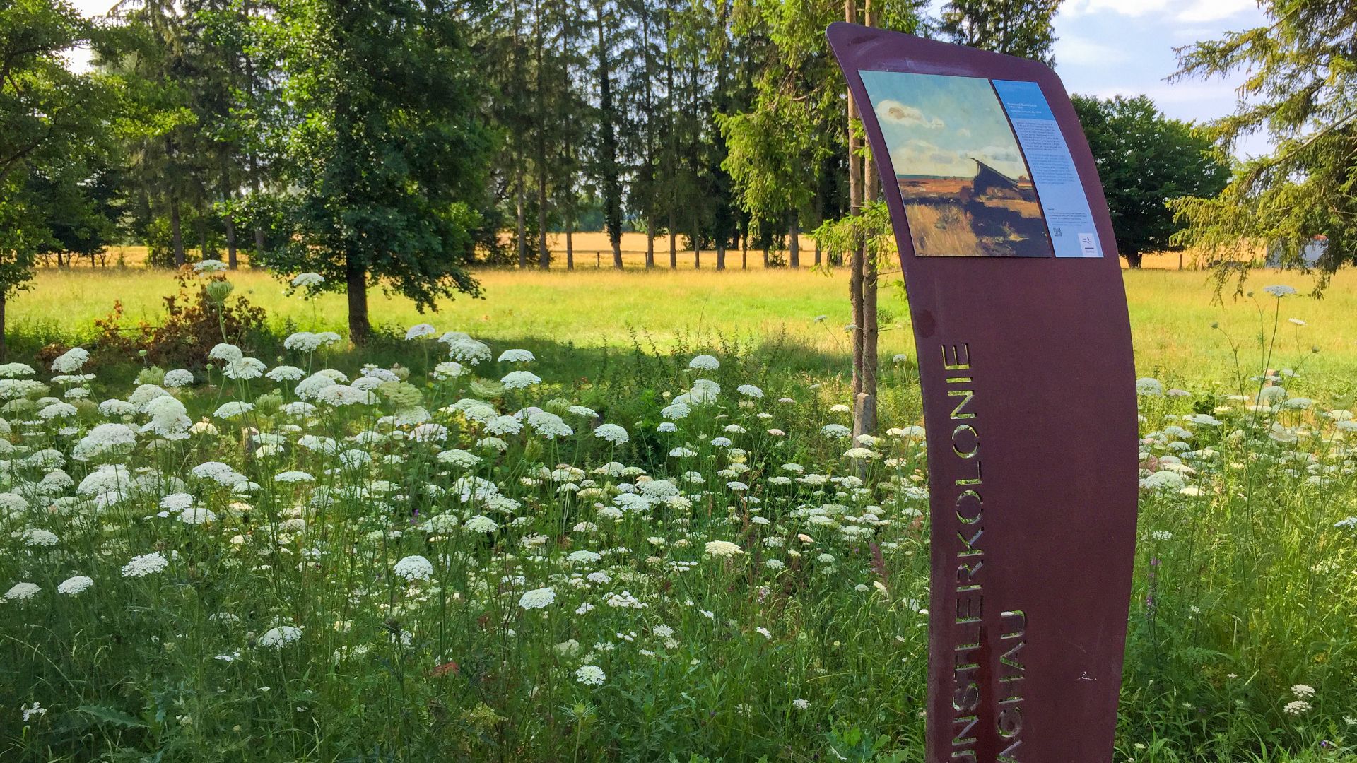 Foto einer Stele des Dachauer Künstlerwegs dahinter Wiese, Büsche und Bäume.