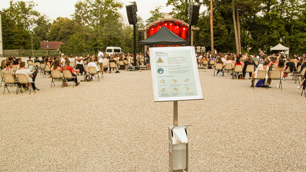 Openairkonzertbestuhlung mit viel Abstand zwischen den paarweise aufgestellten Stühlen, davor die Hygieneregelübersicht auf einem Schild