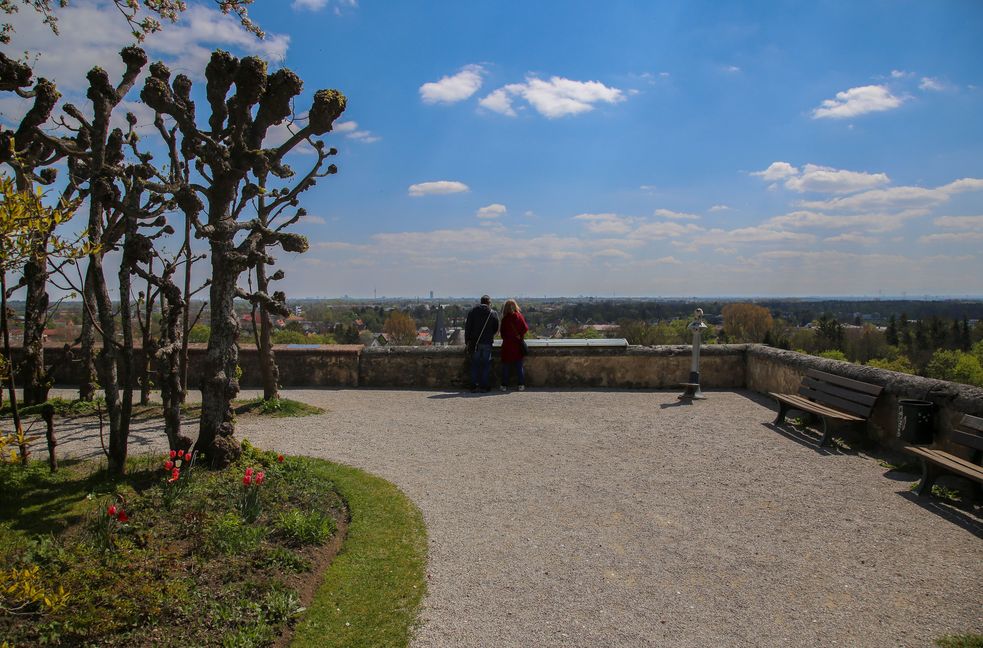 Foto von einem Kiesweg im Hofgarten Richtung Schlossmauer, wo zwei Besucher vor weiß-blauem Frühlingshimmel die Aussicht genießen.