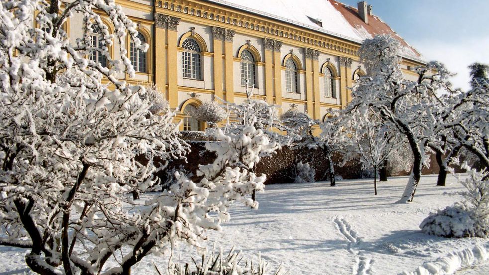 Verschneiter Hofgarten, auf den Bäumen und Sträuchern liegt eine dicke Schneedecke. Das Dach von Schloss Dachau ist mit Schnee bedeckt. Die Sonne scheint.