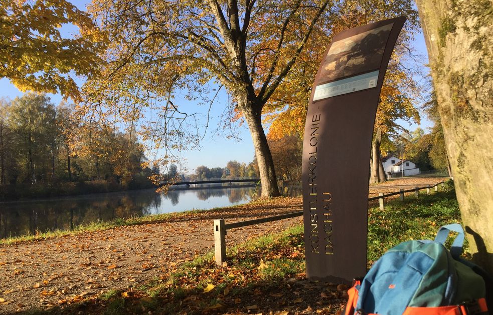 Fotografie einer Stele des Künstlerwegs in Dachau im Herbst, am Baum lehnt ein blauer Rucksack, neben dem Weg fließt die Amper.