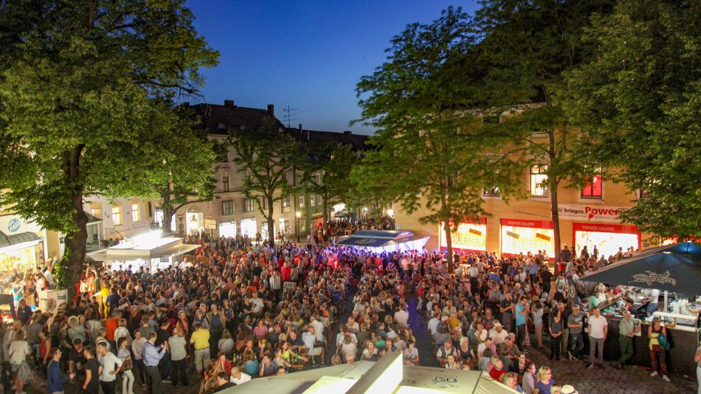 Viele Menschen sitzen und stehen auf einem Platz mit Ständen