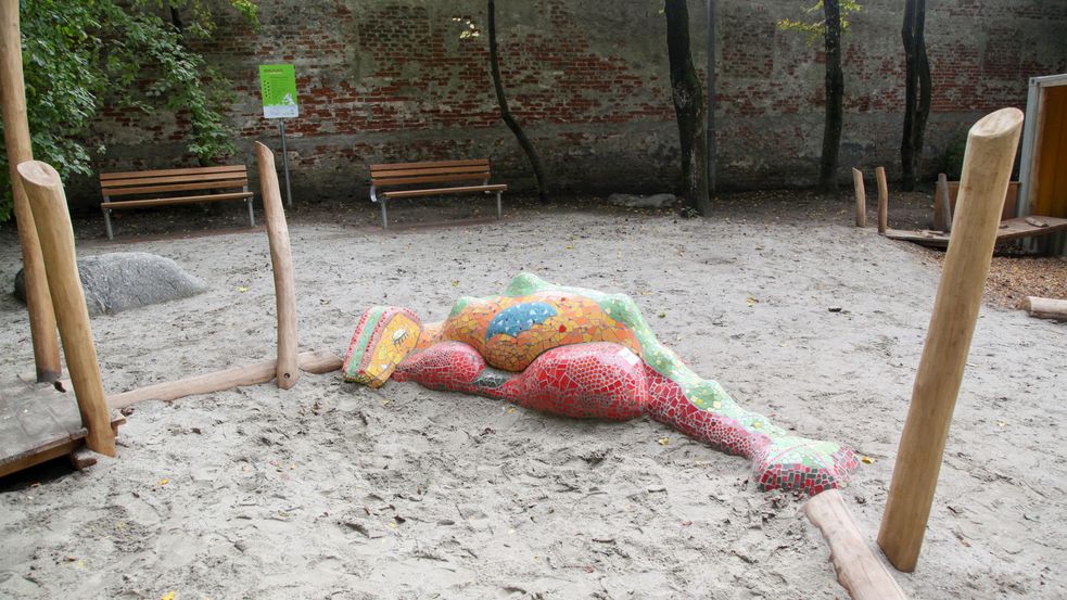 Sandspielbereich mit Mosaikdrachen und Bänke im Hintergrund