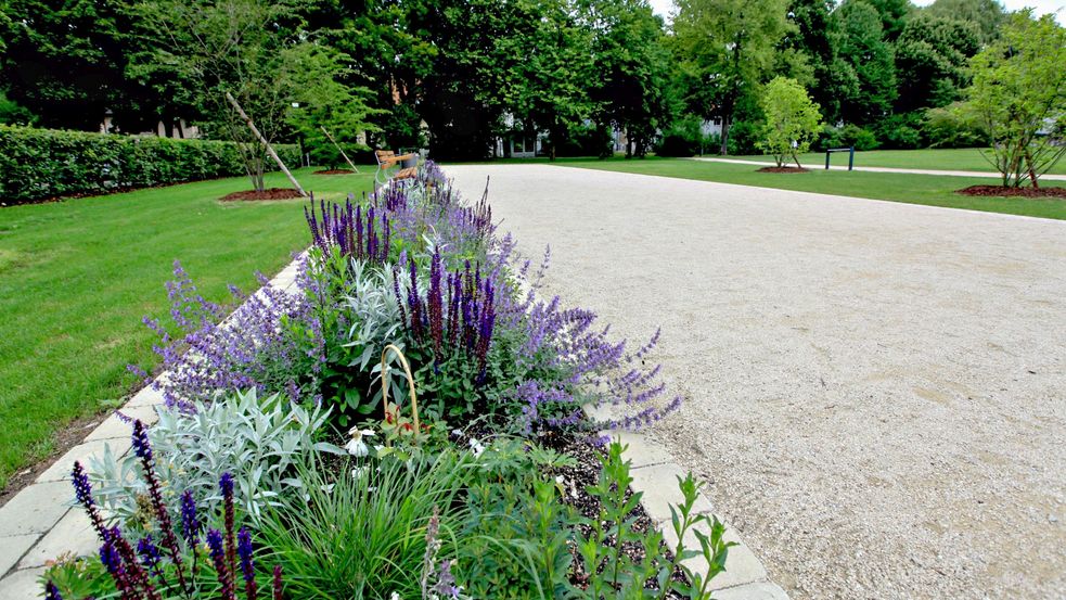 Der Moorbad-Park in Dachau, Blumenrabatten säumen den gekiesten Weg. Große Laubbäume im Hintergrund.