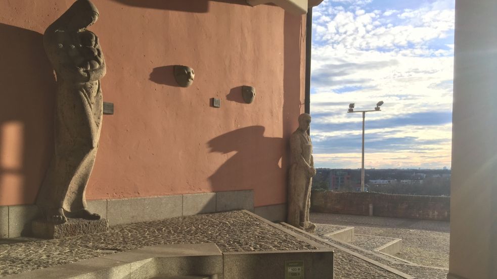 Fotografie in Durchgang zu Dachauer Rathausterrasse, links an der Wand stehen zwei Skulpturen, eine weibliche mit Kind, ein Mann auf ein Werkzeug gestützt. Dahinter öffnet sich der Ausblick nach München.