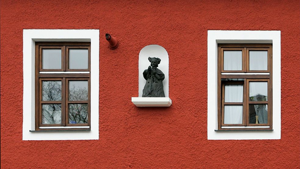 Detailfotografie einer roten Hausfassade mit zwei weiß gerahmten Fenstern. Zwischen den Fenstern ist eine weiß abgesetzte Nische mit einer Skulptur eines Mädchens in Dachauer Tracht.