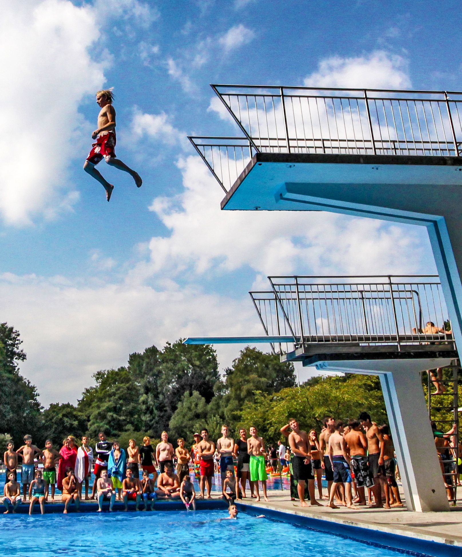 Junge springt vom 5 Meterturm im Freibad, vom Beckenrand schauen ihm einige Jugendliche dabei zu