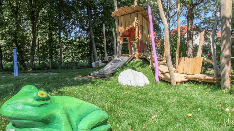 Holzfroschskulptur auf grüner Wiese, Rutsche im Hintergrund