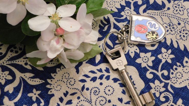 Weiße Blumen und Zimmerschlüssel mit Schlüsselanhänger "Dachauer Stadtwappen" auf blau-weißer Tischdecke