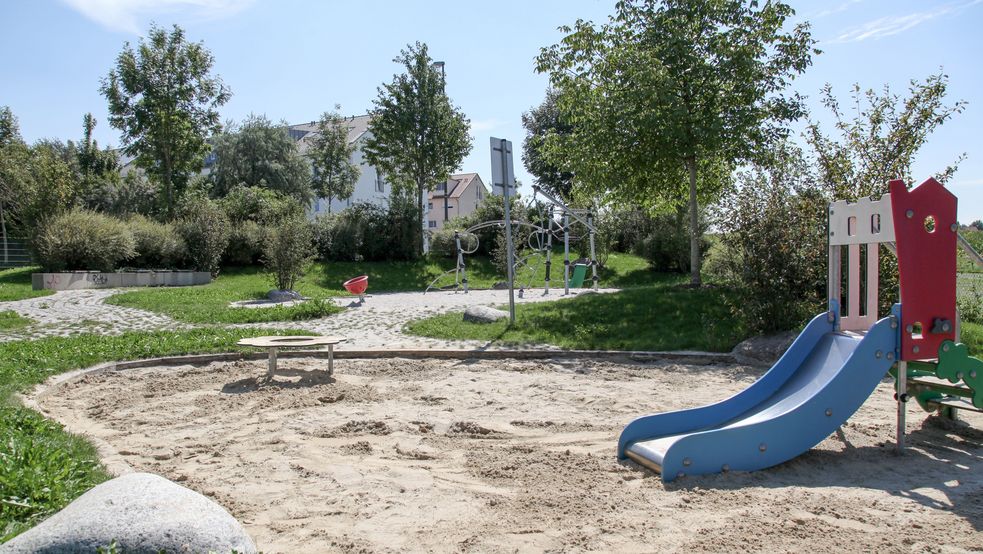 Sandkasten mit kleiner Rutsche, umgeben von Bäumen und Wiese