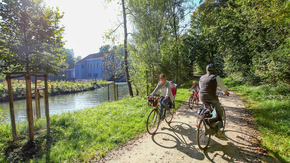 Radlfahrende auf Radweg neben grünem Flussbett