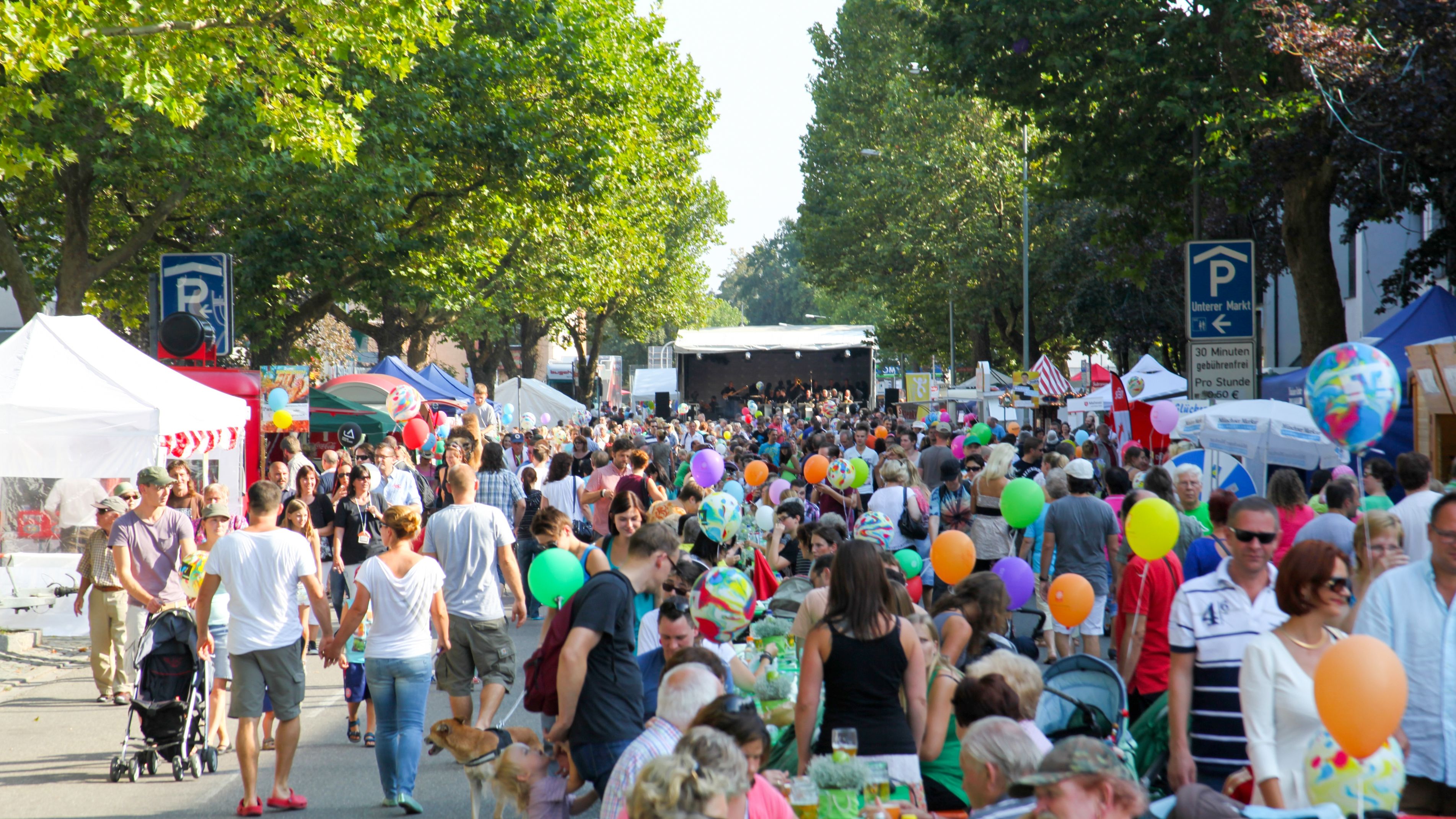 Viele Menschen laufen oder sitzen gemeinsam auf einer langen Straße mit Festivalständen zu beiden Seiten