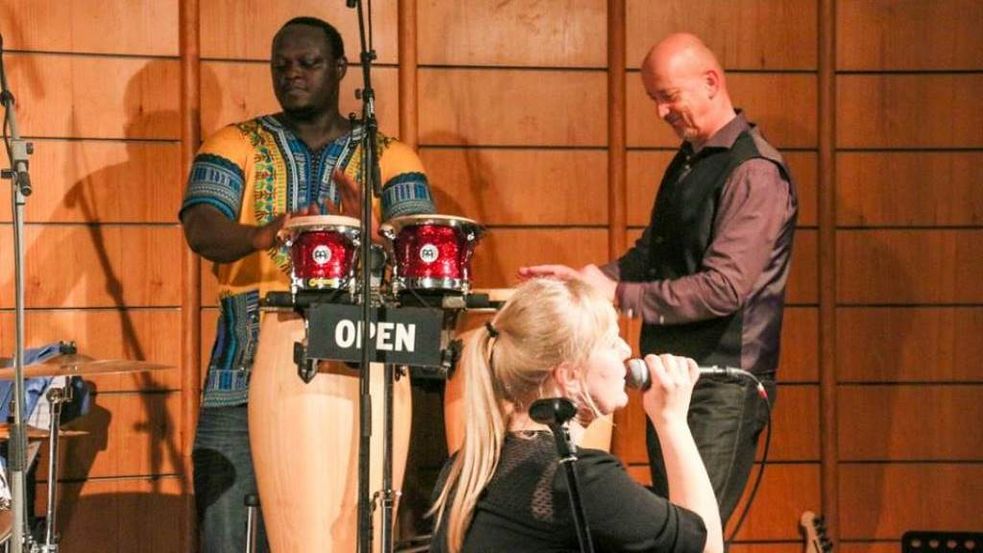 Sängerin mit Mikrofon, im Hintergrund zwei Musiker an Trommeln