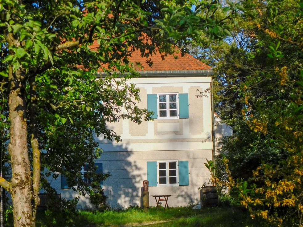 Fotografie der sogenannten "alten Moosschweige" in Dachau. Durch den üppig grünen Garten hindurch sieht man das kleine Haus mit blauen Fensterläden.