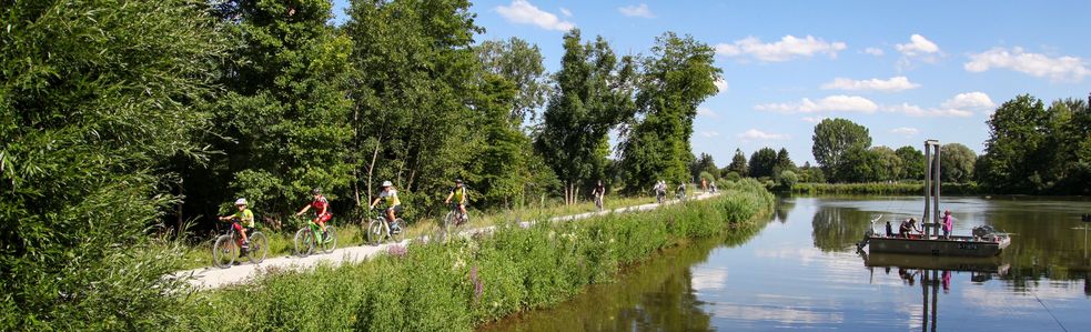 Radlfahrer fahren auf dem Radweg neben der Amper auf der eine Fähre vorbeifährt