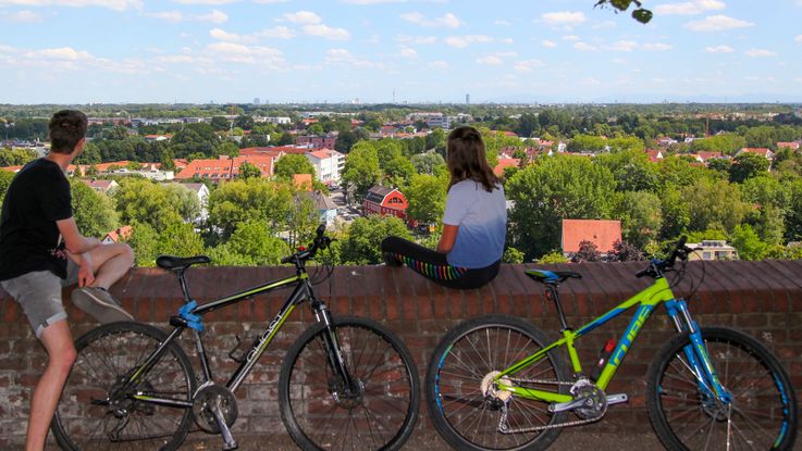 Zwei Radfahrer genießen auf der Schlossmauer sitzend den Ausblick nach München